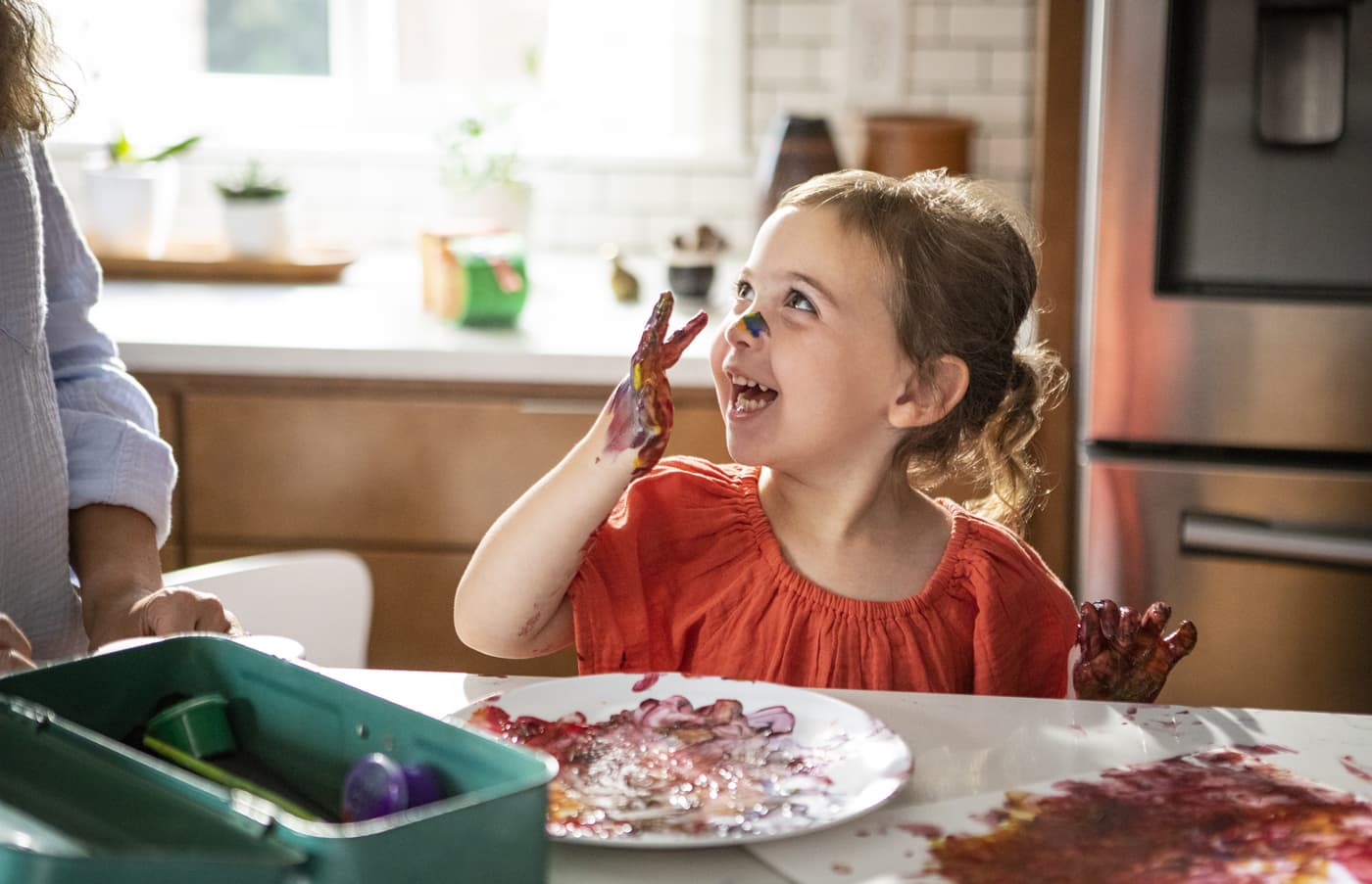 A little girl with paint on her hands and face looking at her mom