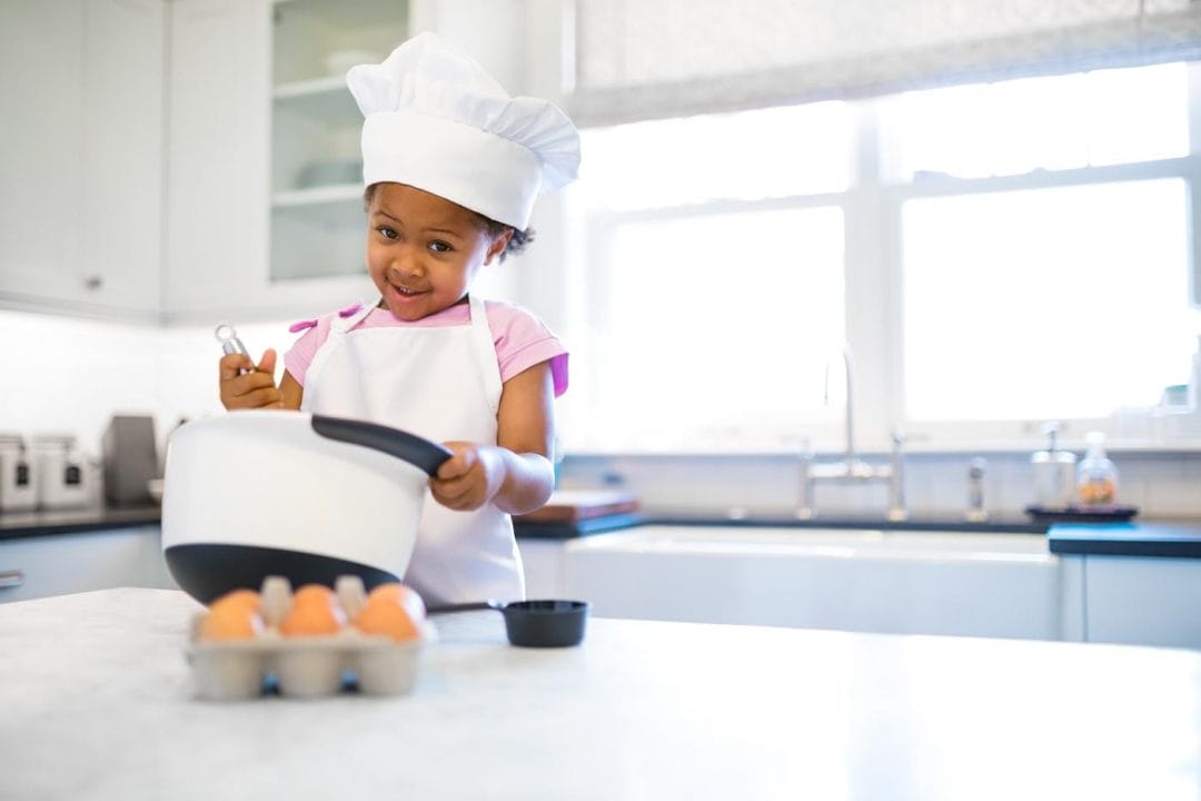 A little girl in a chefs hat and apron mixing something in a bowl with eggs on a kitchen counter